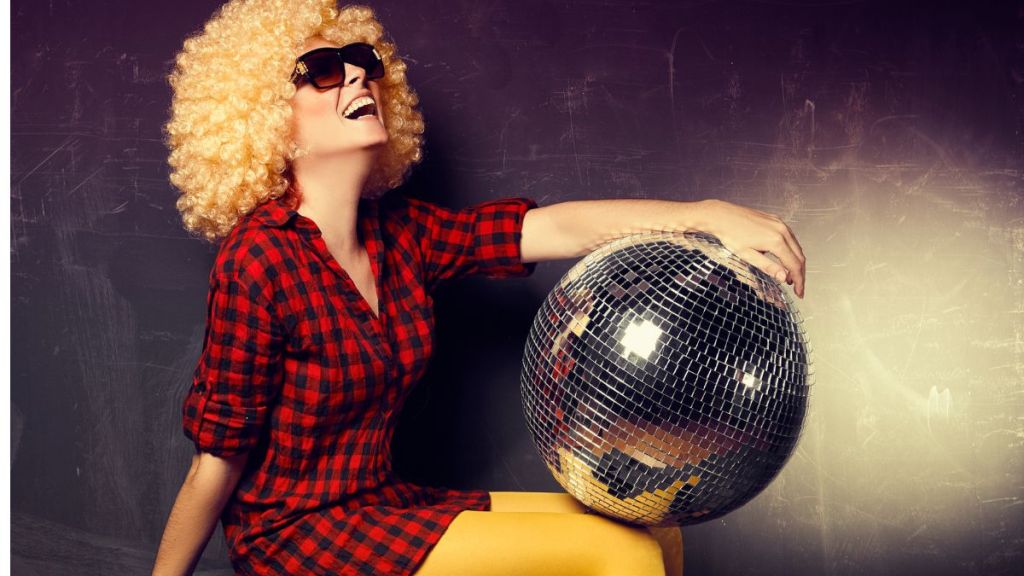 A woman in a large curly blonde wig holding a disco ball on her lap and laughing with her mouth wide open