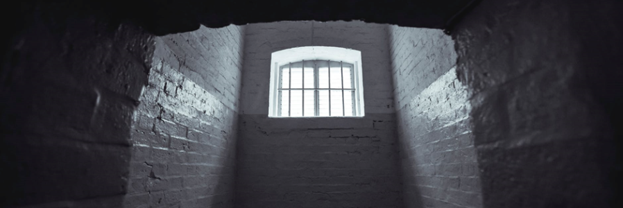 The black and white image of the inside of a prison cell with a small widow with bars through which daylight is streaming